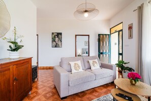 A living room with a sofa, a wooden cabinet, a table with a vase, and a hanging light fixture. - GuestReady - Quinta A Lealdade (Arco de São Jorge)