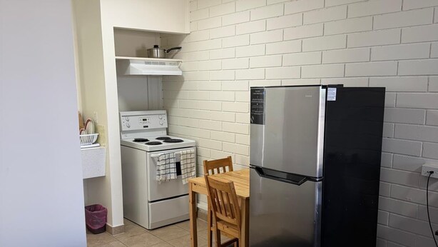 A compact kitchenette with a stove, oven, and refrigerator, a small wooden table, and a bed in the foreground. - Peacevillamotel (Dawson Creek)