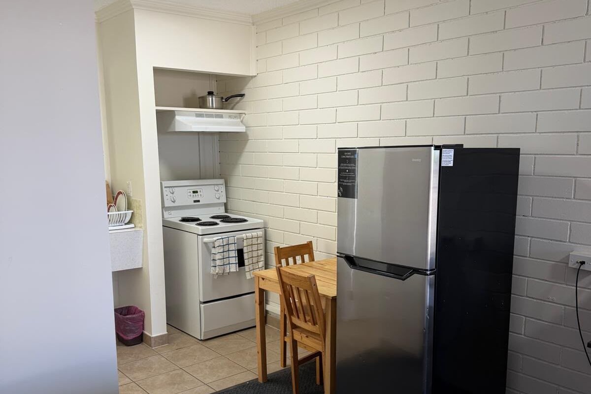 Une petite cuisine équipée avec une cuisinière, un four et un réfrigérateur, une petite table en bois et un lit au premier plan.