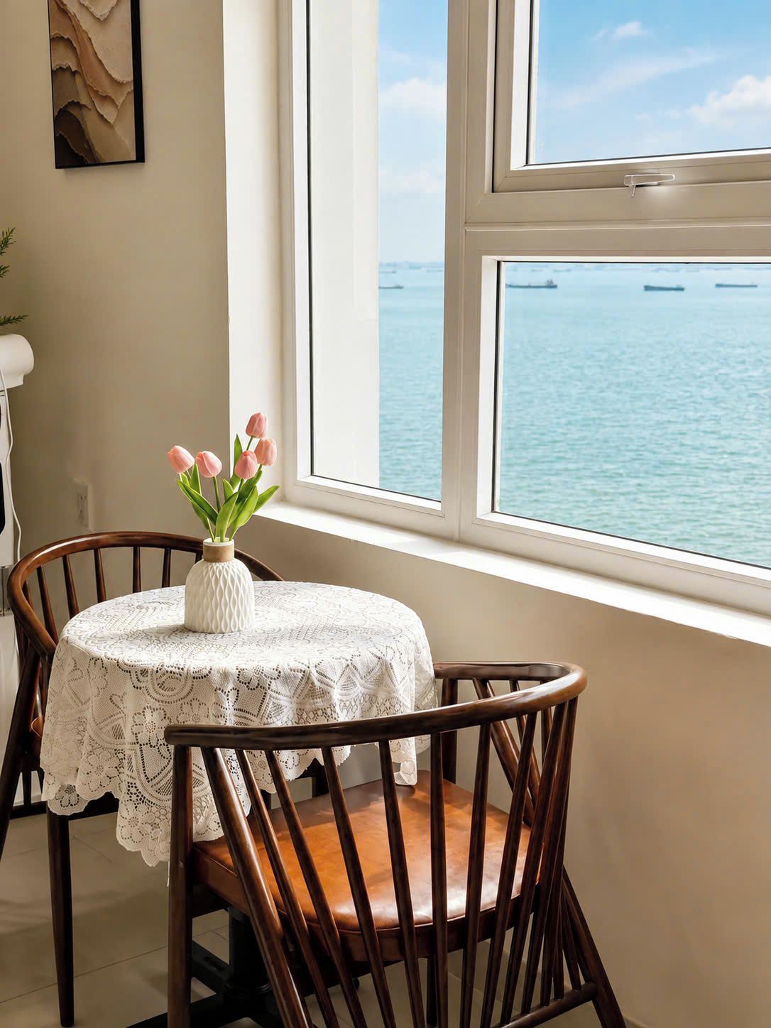 A dining area with a table set for two, a vase with flowers, and a view of the sea through a window.