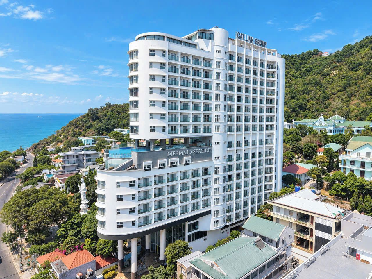 A multi-story hotel with a curved facade, named 'Mermaid Seaside,' located near a coastline with a view of the sea.