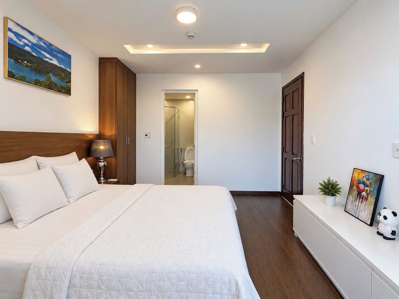 A hotel room with a bed, bedside lamp, a small plant, a framed picture, and a door leading to a bathroom.