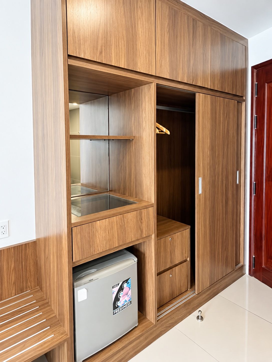 A wooden wardrobe with multiple compartments, a small refrigerator, and a hanging space.