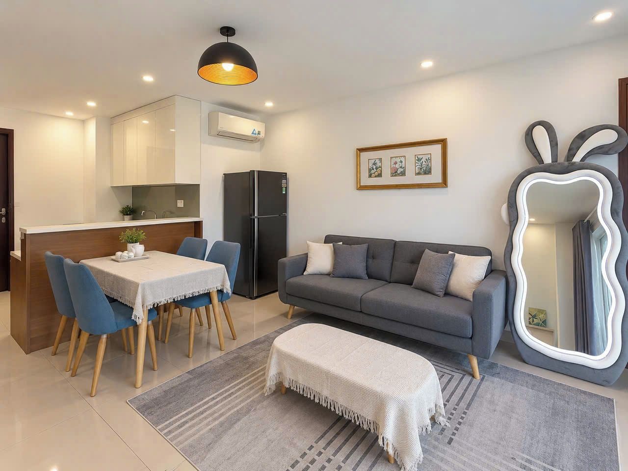 A modern living room with a grey sofa, a dining table with chairs, a kitchen area with an air conditioner, and a unique mirror decoration.
