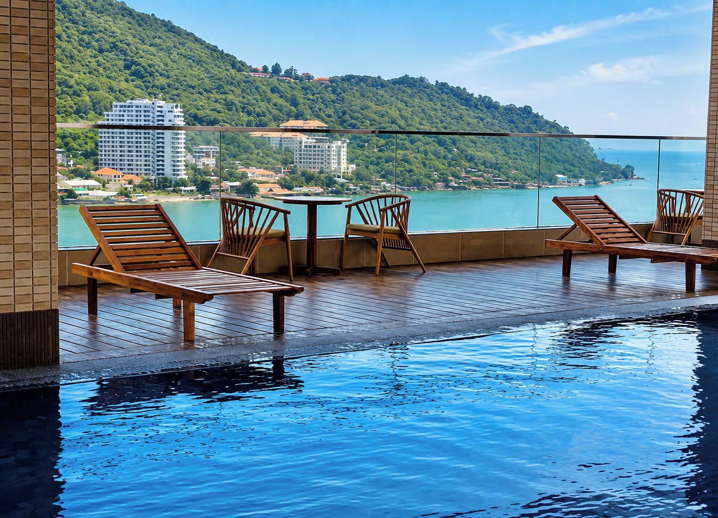 A pool area with wooden loungers and a table, overlooking a body of water and buildings.