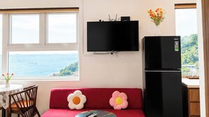 A compact living space with a sofa, a small round table, a television mounted above the sofa, a refrigerator, and a view of the sea through a window. - Moc An Collection - Mermaid Seaside VT (Vung Tau)