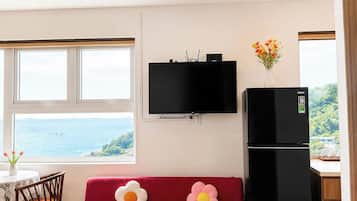 Un espacio habitable compacto con un sofá, una pequeña mesa redonda, un televisor sobre el sofá, un refrigerador y vista al mar por una ventana.