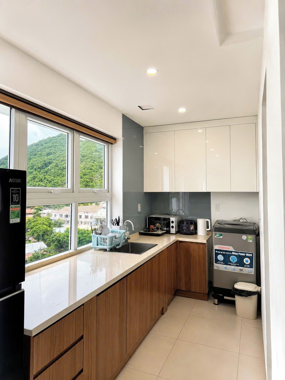 Una cocina moderna con refrigerador, microondas y cafetera, una ventana con vista a la vegetación y un bote de basura.
