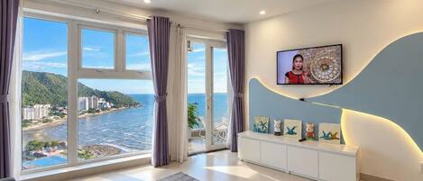 A modern living room with a large window overlooking a coastal view, a white entertainment unit, and a mounted television.
