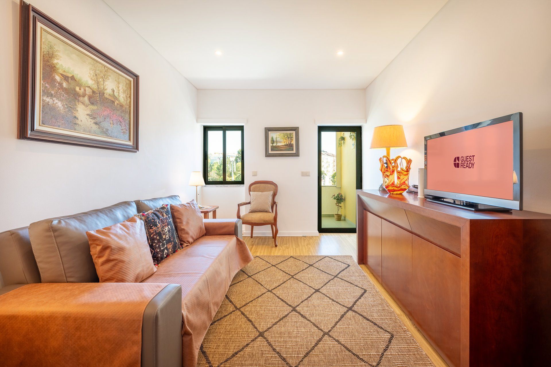 Uma sala de estar moderna com um sofá de couro, uma televisão exibindo 'GUEST READY' e um armário de madeira.