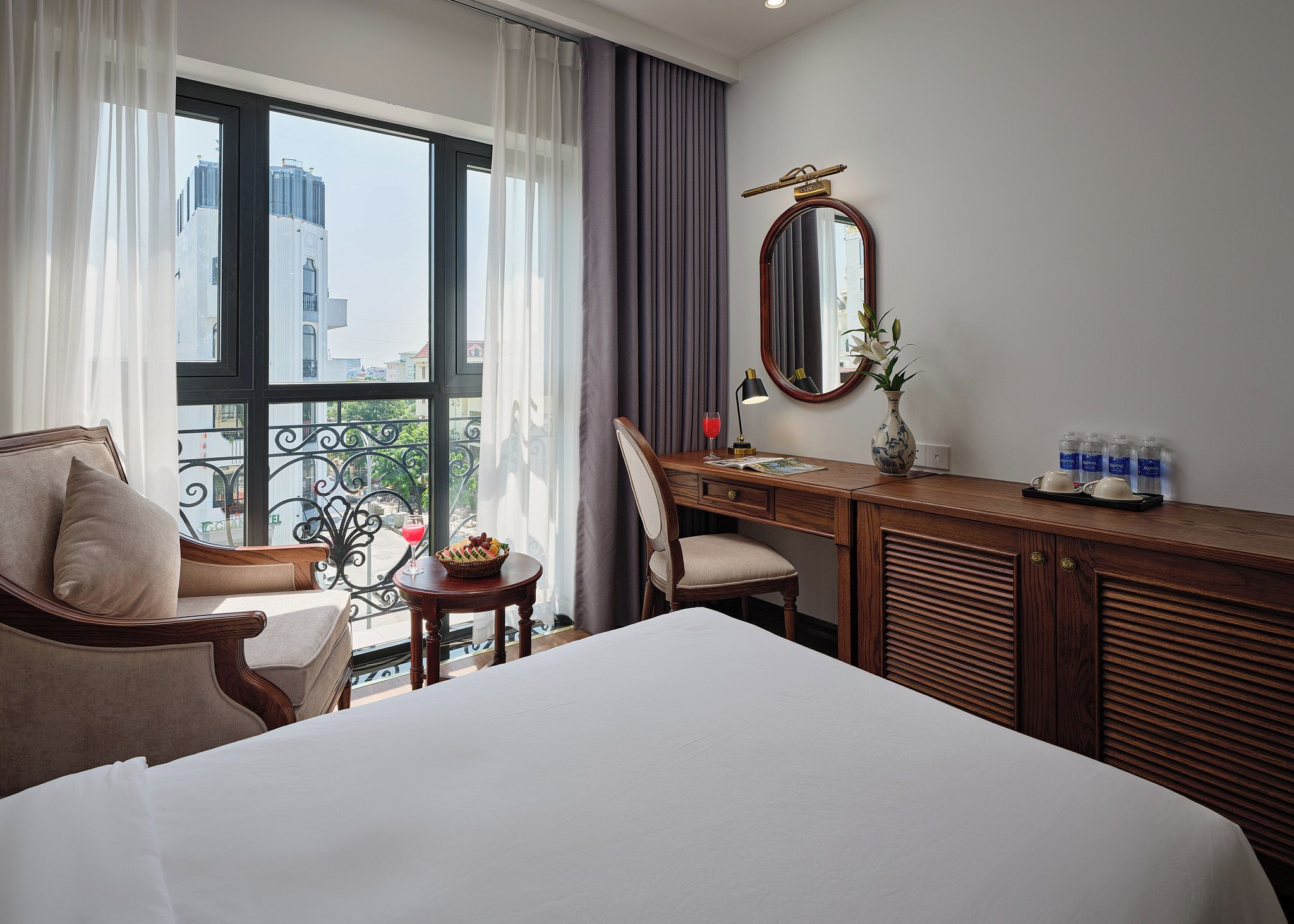 A hotel room with a bed, a desk, a chair, a window with curtains, a mirror, and a vase with flowers.