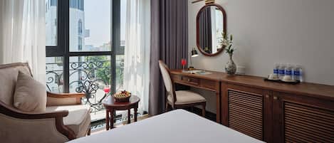 A hotel room with a bed, a desk, a chair, a window with curtains, a mirror, and a vase with flowers.