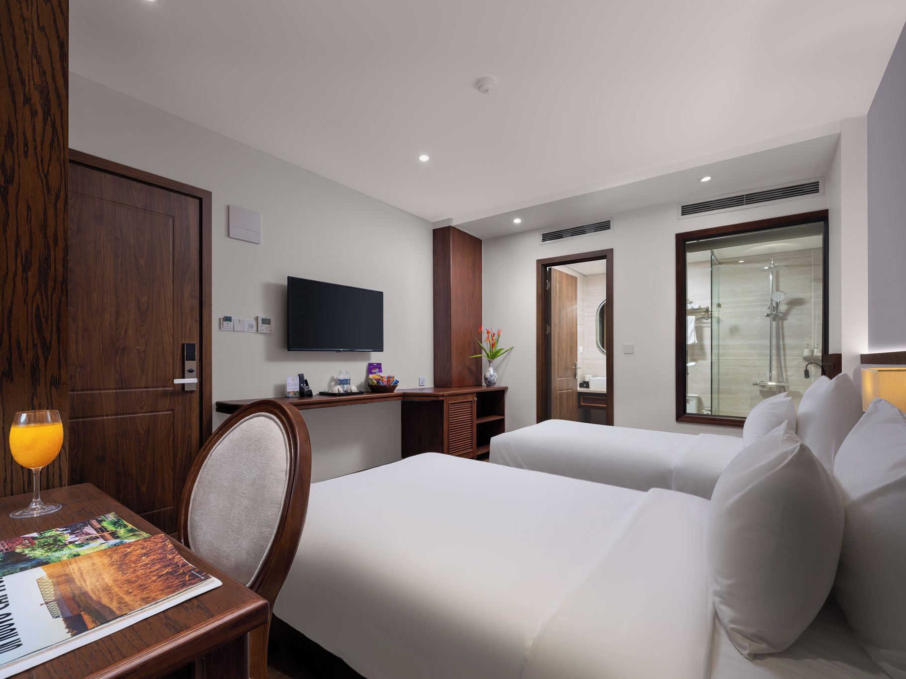Habitación de hotel con dos camas, un televisor, un escritorio y un vaso de jugo de naranja, y un baño visible a través de una puerta abierta.