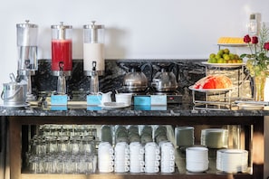 Una barra de desayuno con varios dispensadores de bebidas, una máquina de café y una presentación de frutas.