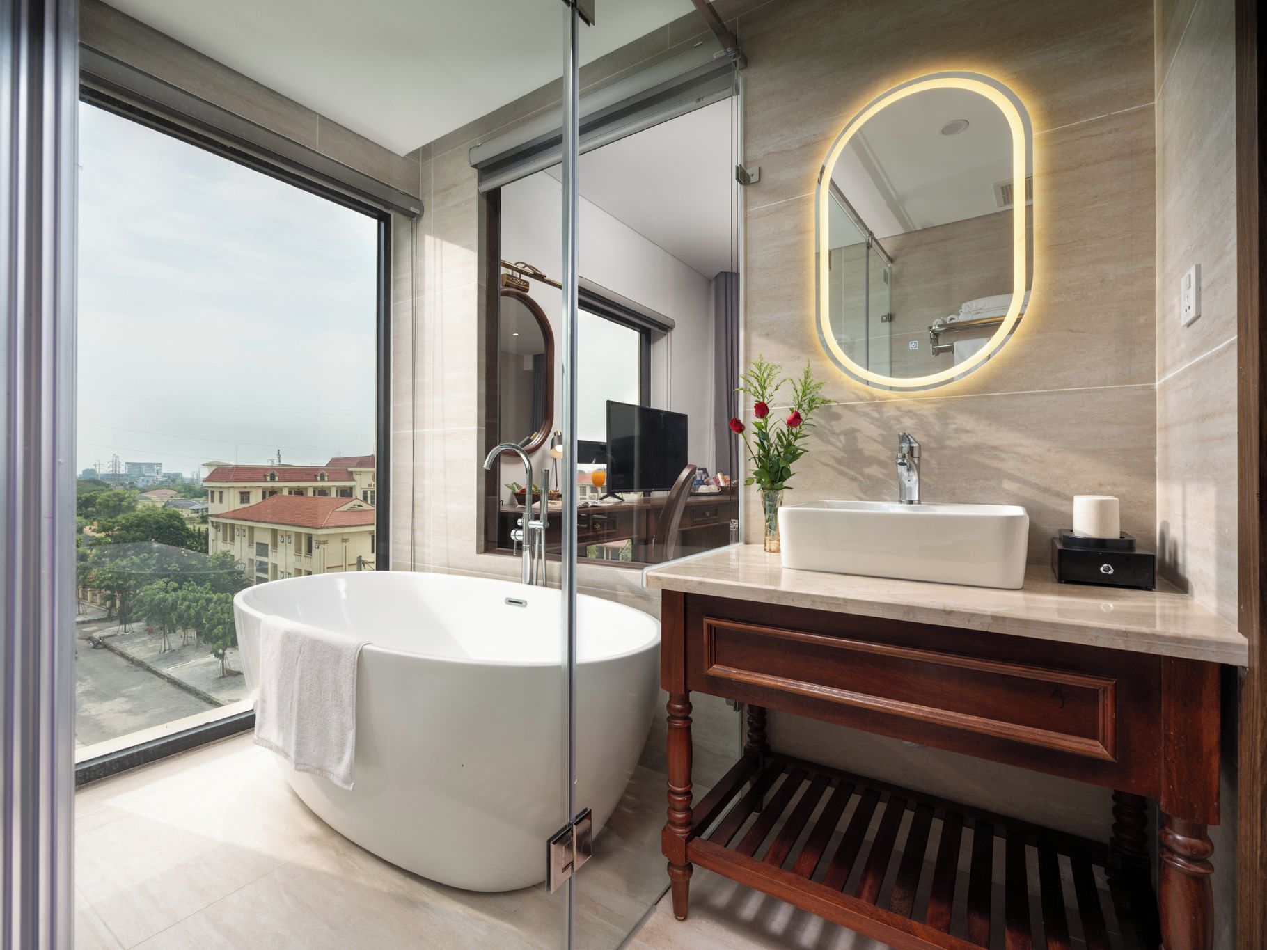 Un baño moderno con una bañera independiente, un tocador con espejo y vistas a la ciudad a través de amplios ventanales.