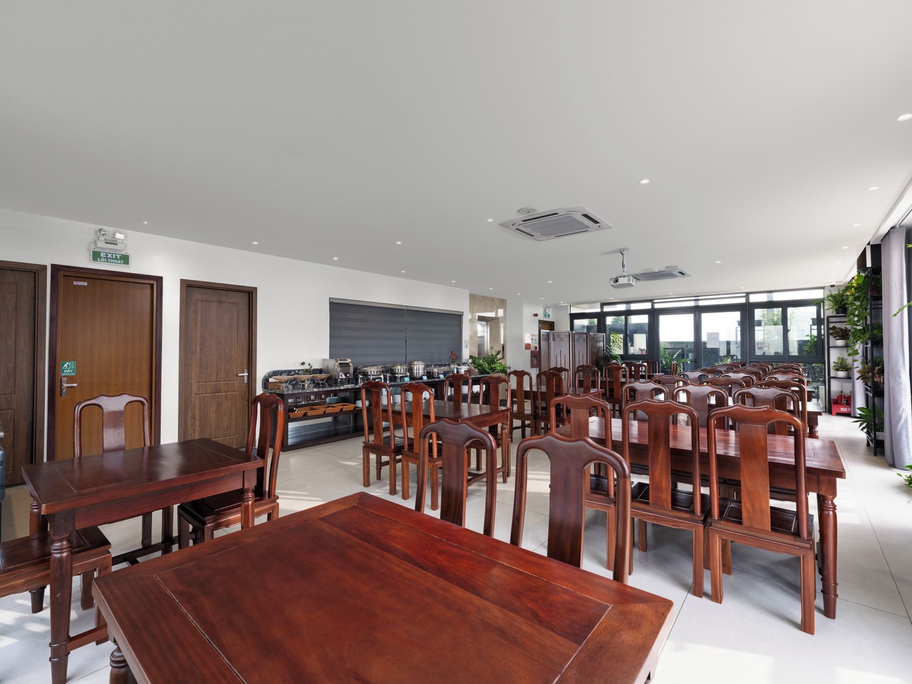 A spacious dining area with multiple wooden tables and chairs, a kitchen in the background, and large windows providing natural light.