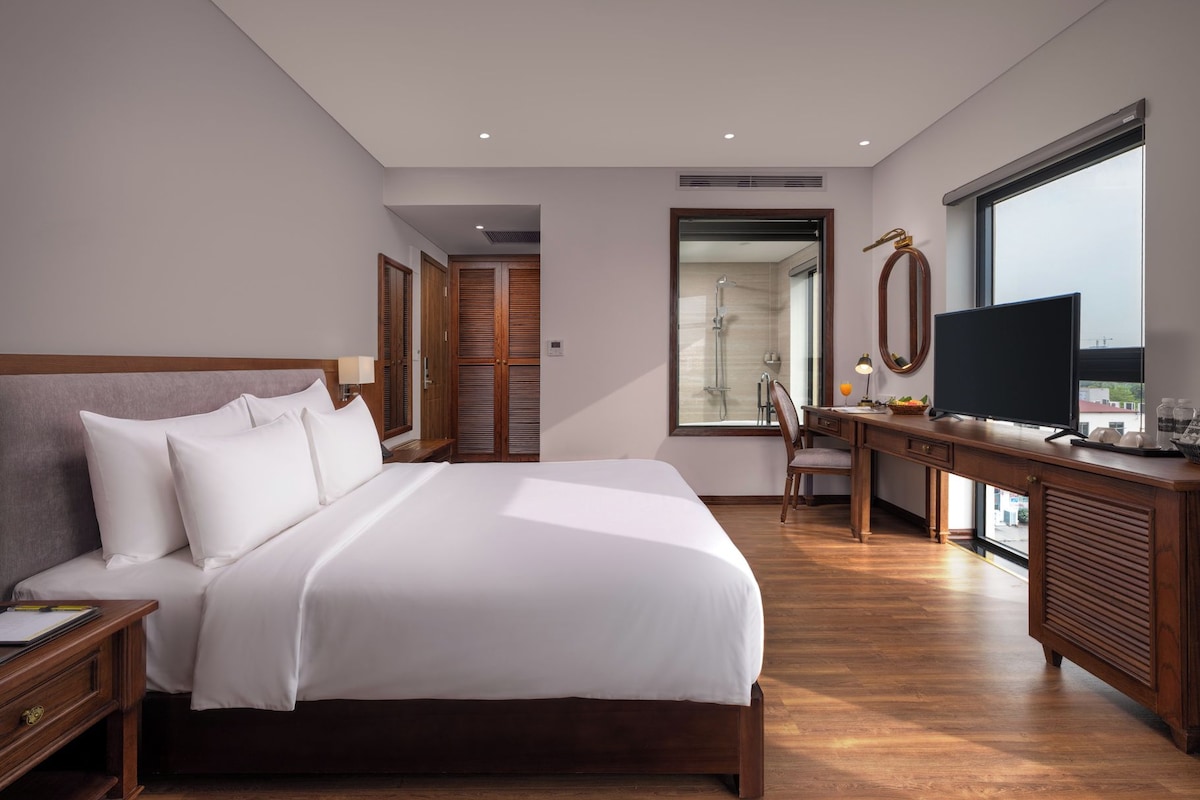 Una habitación de hotel moderna con una cama grande, un escritorio con televisor y vistas amplias por una ventana grande.