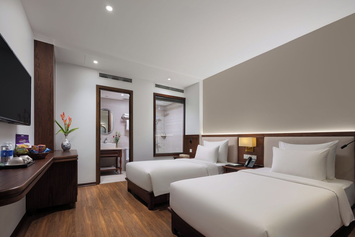 Habitación de hotel con dos camas, televisión, escritorio con un florero y baño visible a través de una puerta abierta.