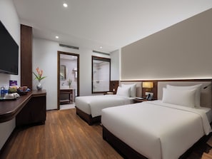 Habitación de hotel con dos camas, televisión, escritorio con un florero y baño visible a través de una puerta abierta.