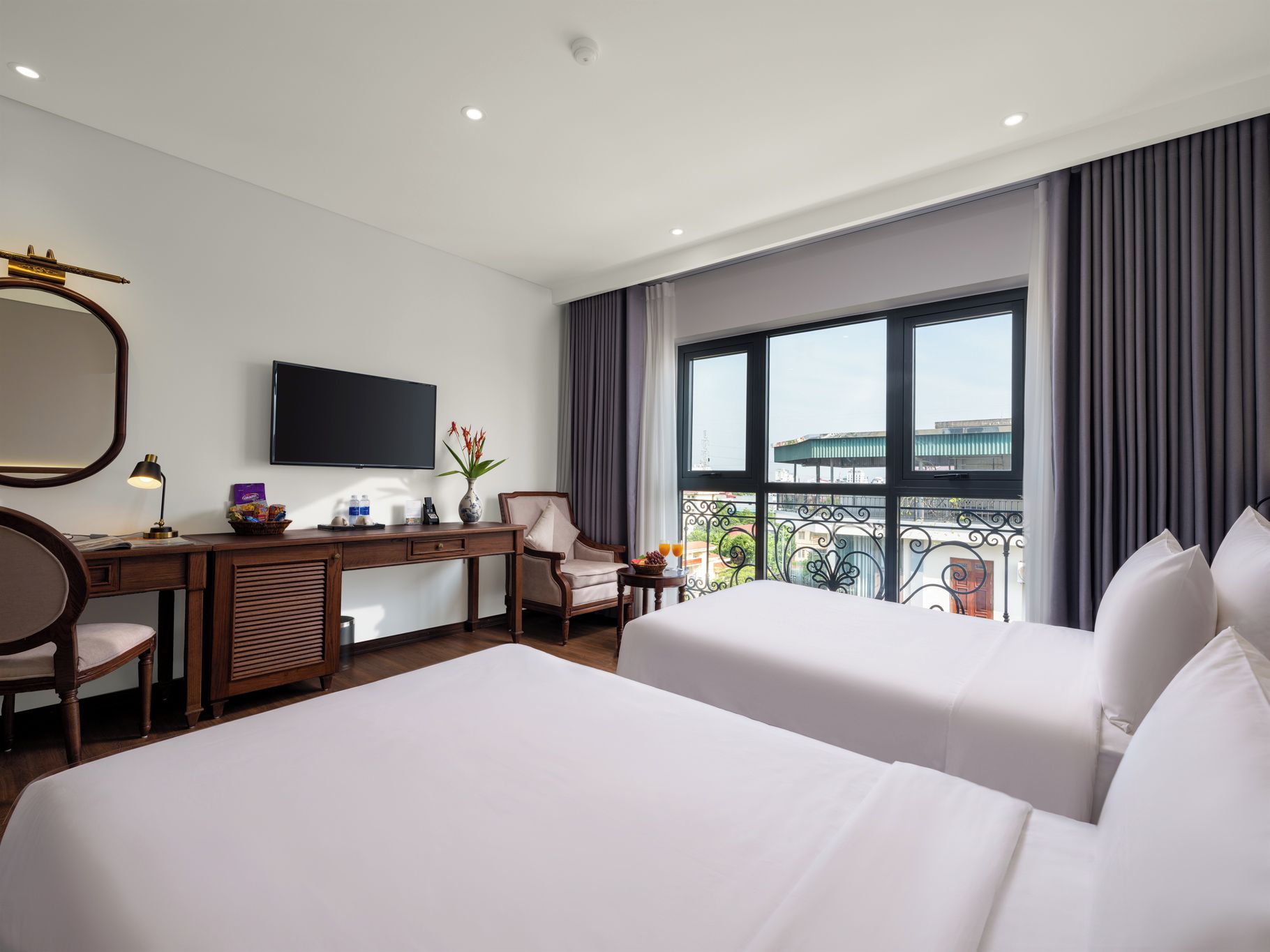 A hotel room with two beds, a desk, a chair, a television, and a balcony with a view of buildings.