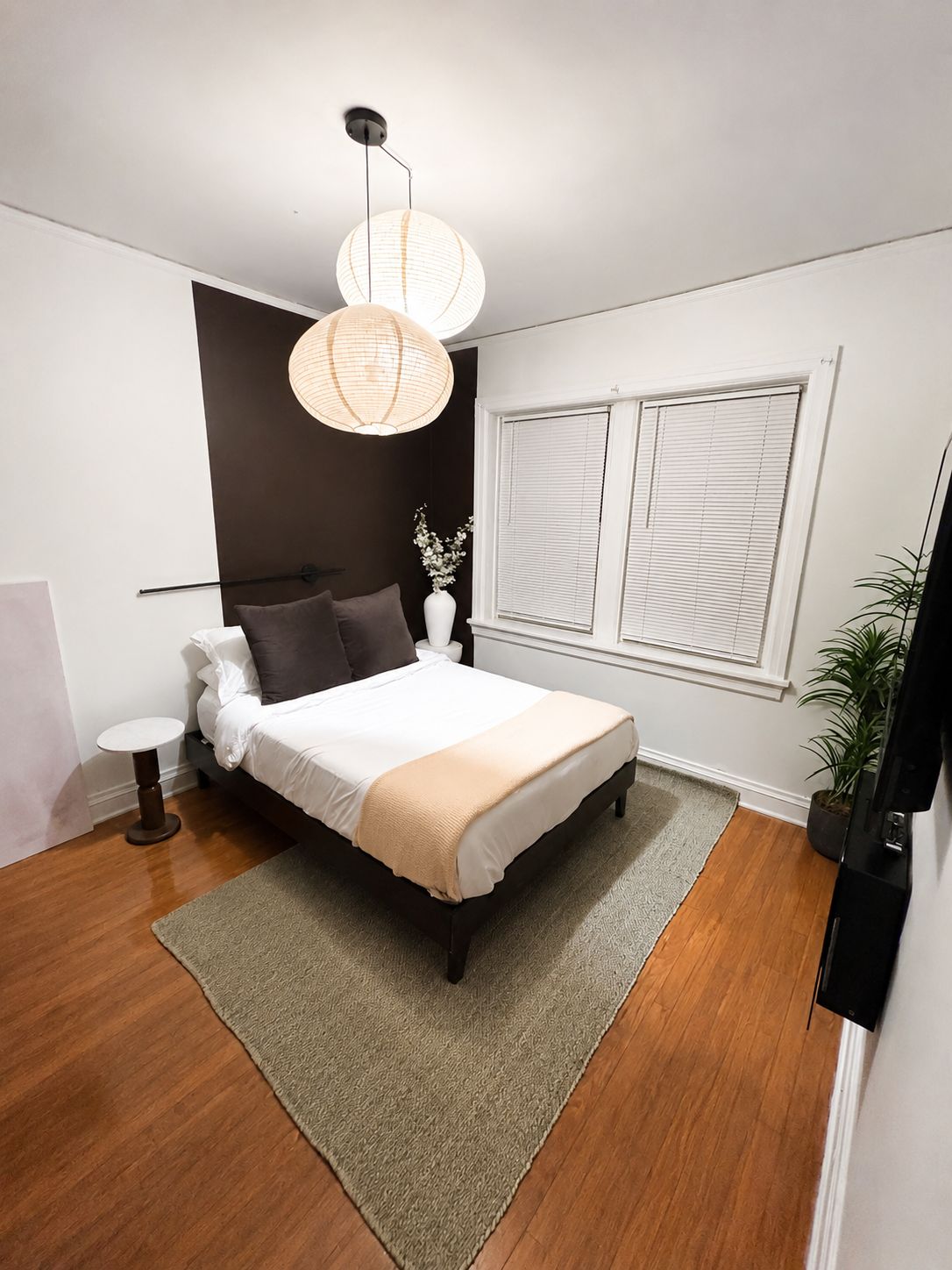 A bedroom with a bed, bedside tables, a window with blinds, a hanging light fixture, and a potted plant.