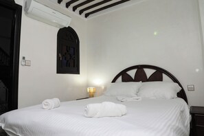 Una camera d'albergo con un letto, un condizionatore d'aria a muro, una porta con oblò e un comodino con lampada.