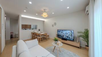 A modern living room with a sofa, a dining area, a flat-screen TV, and a kitchen in the background.