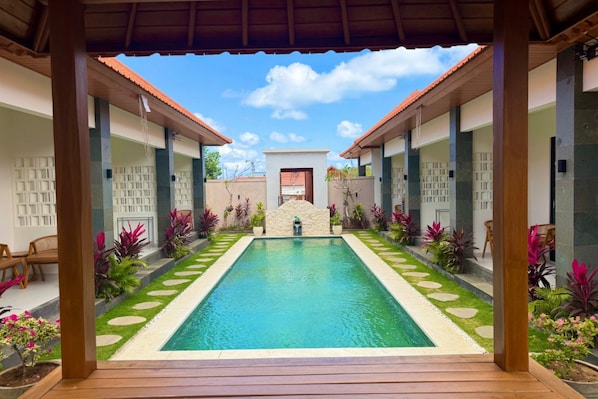 A swimming pool with a stone pathway leading to a covered area, surrounded by potted plants and a wooden deck. - Merta Landuh Bali (Kutuh)