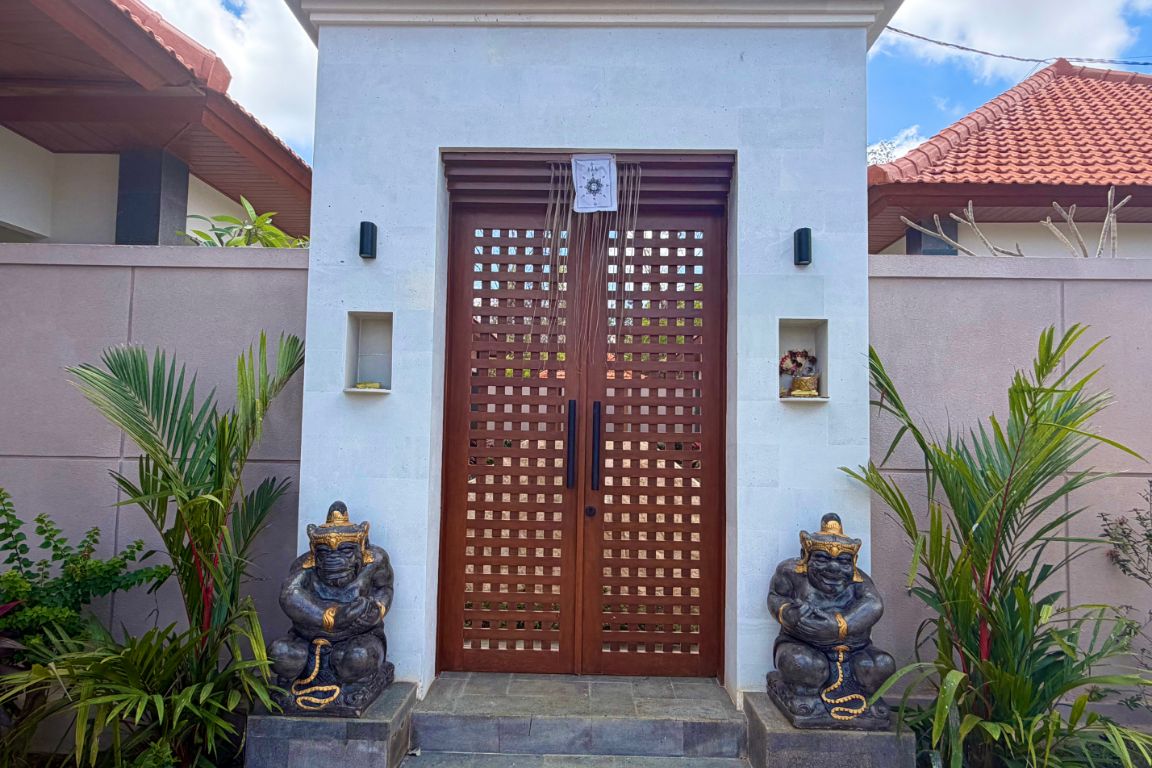 Entrada de edificio con puerta de madera, dos estatuas y un pequeño estante con una flor.