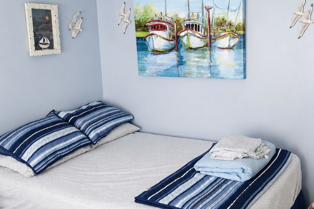 Een slaapkamer met een bed, kussens, een schilderij van boten en vogelfiguurtjes aan de muur.