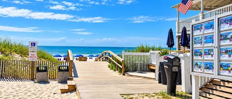 A wooden boardwalk leading to a beach with a no pets sign, a trash can, and a display of property listings.
