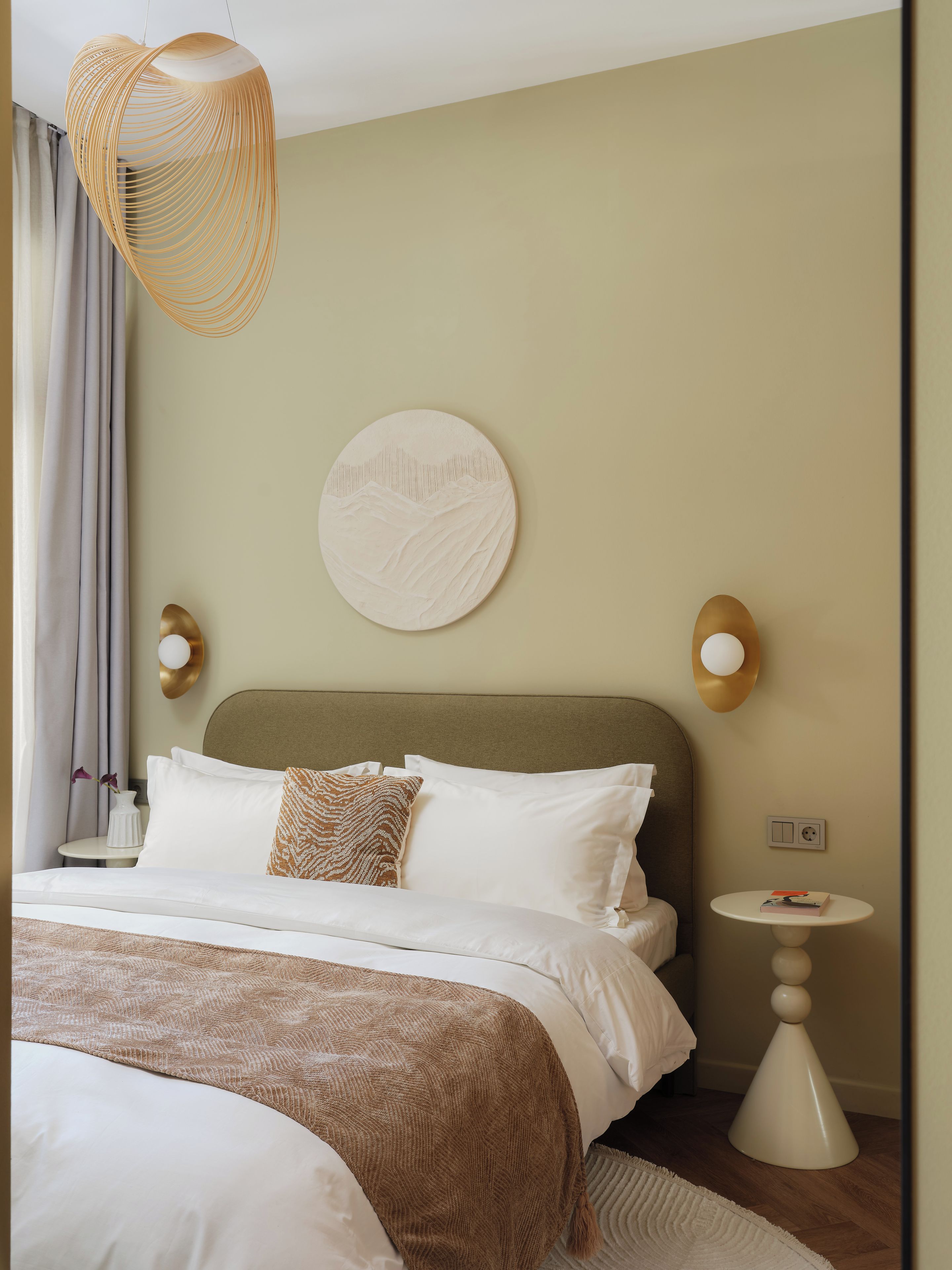 Ein Hotelzimmer mit einem Bett, einer runden Wanddekoration, zwei Wandlampen, einem Nachttisch mit einem Buch und einer Hängeleuchte.