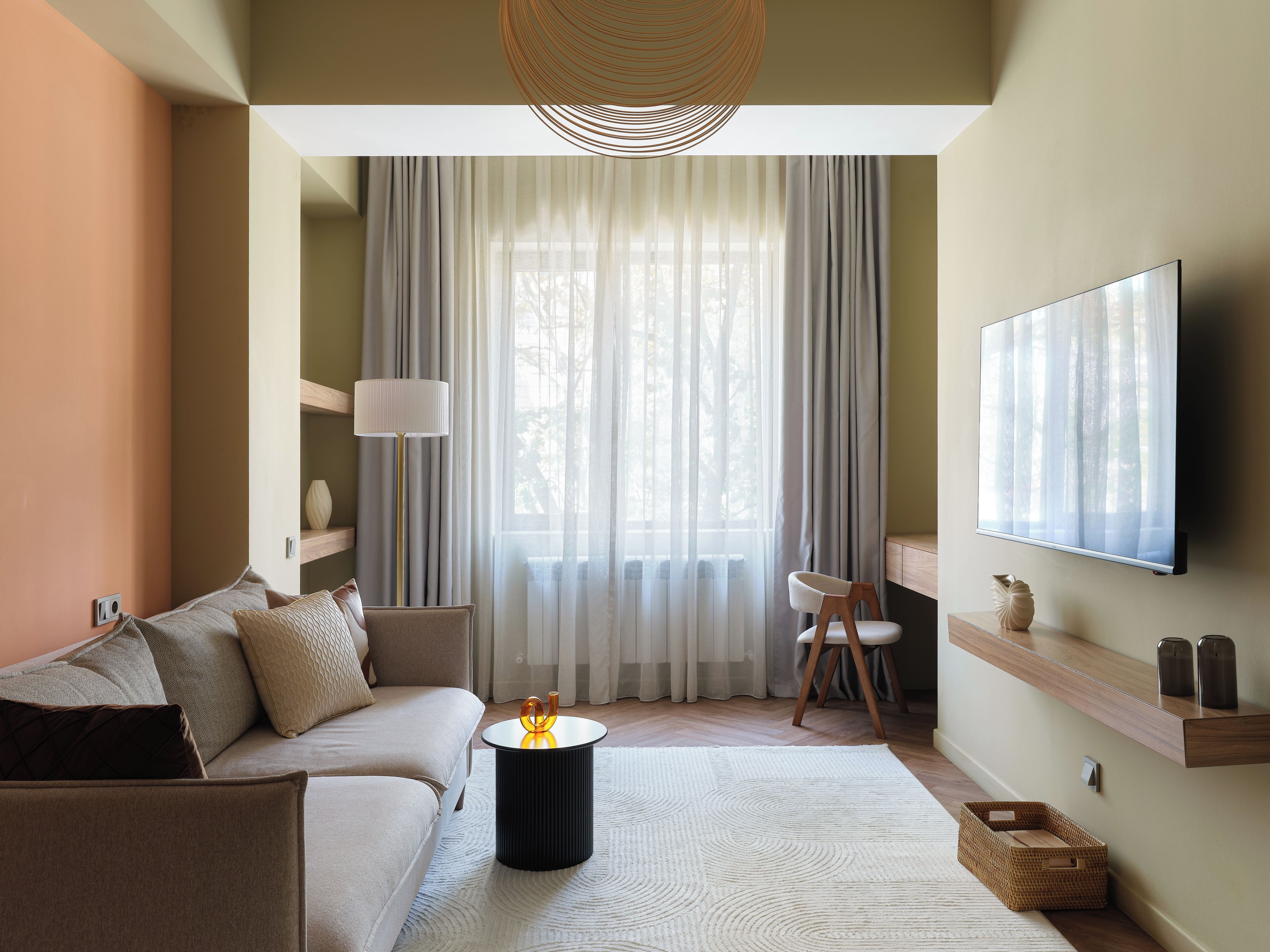 A modern living room with a sofa, a coffee table, a TV mounted on the wall, and a large window with sheer curtains.