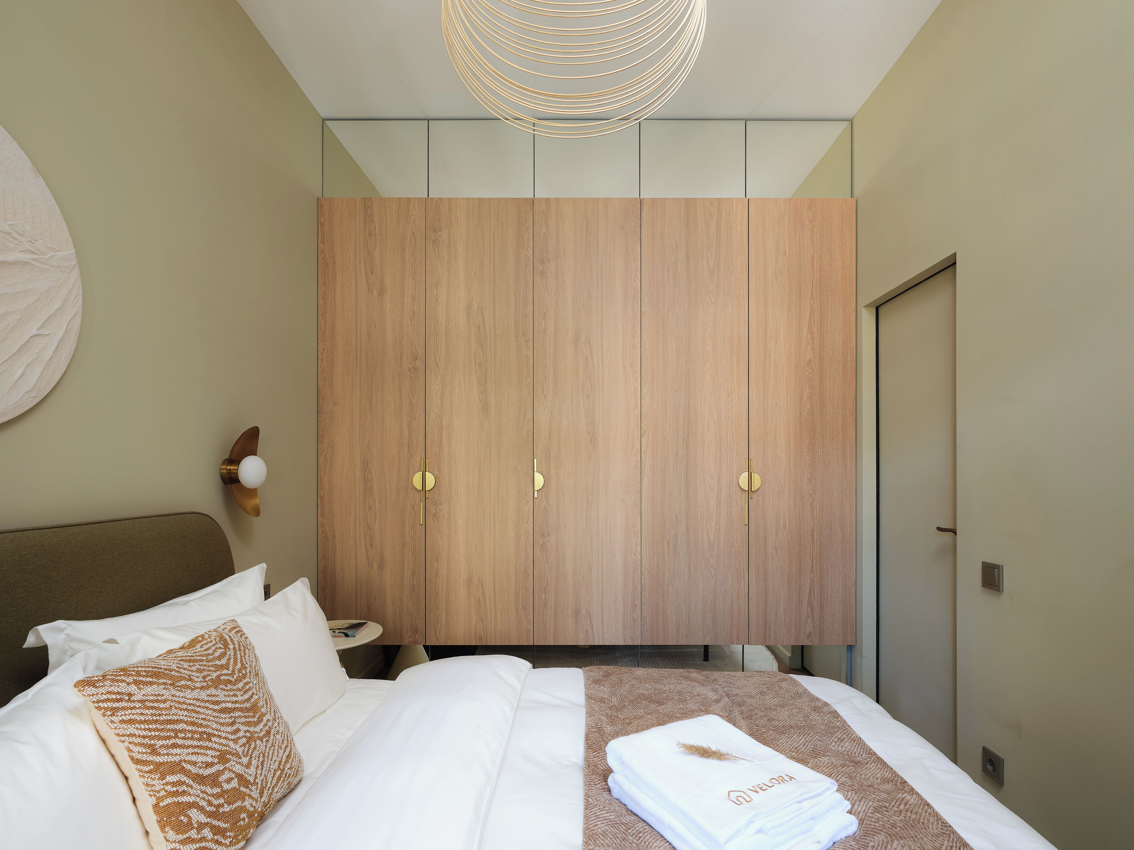 A hotel room with a bed, a wooden wardrobe, a round light fixture, and a wall-mounted round mirror.