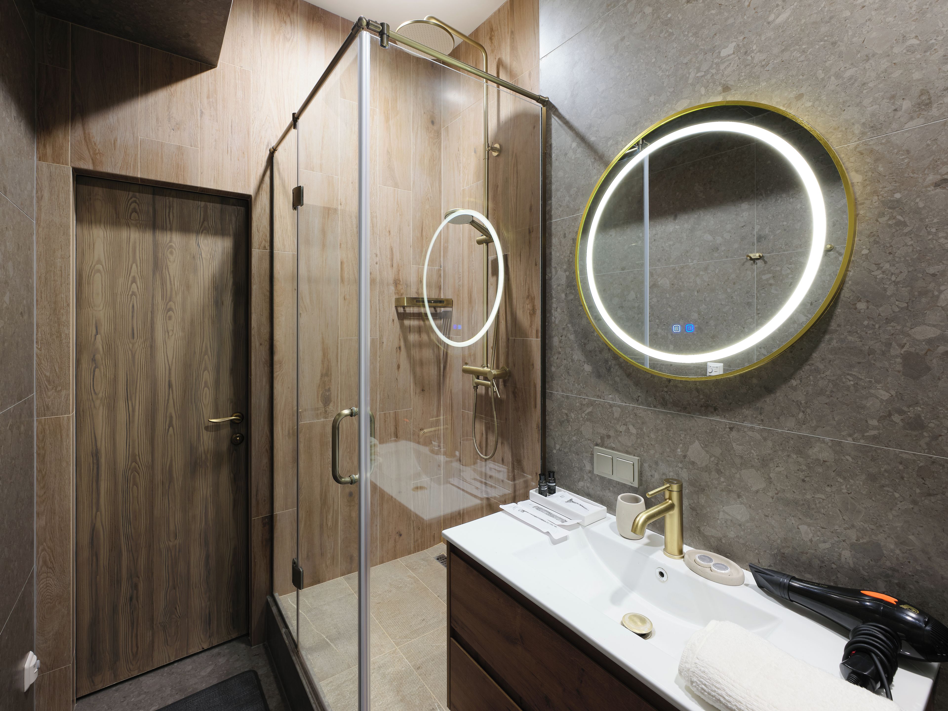 Une salle de bain moderne avec une douche à l’italienne en verre, un grand miroir rond avec éclairage intégré et un lavabo blanc doté d’un robinet doré.
