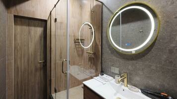 Une salle de bain moderne avec une douche à l’italienne en verre, un grand miroir rond avec éclairage intégré et un lavabo blanc doté d’un robinet doré.