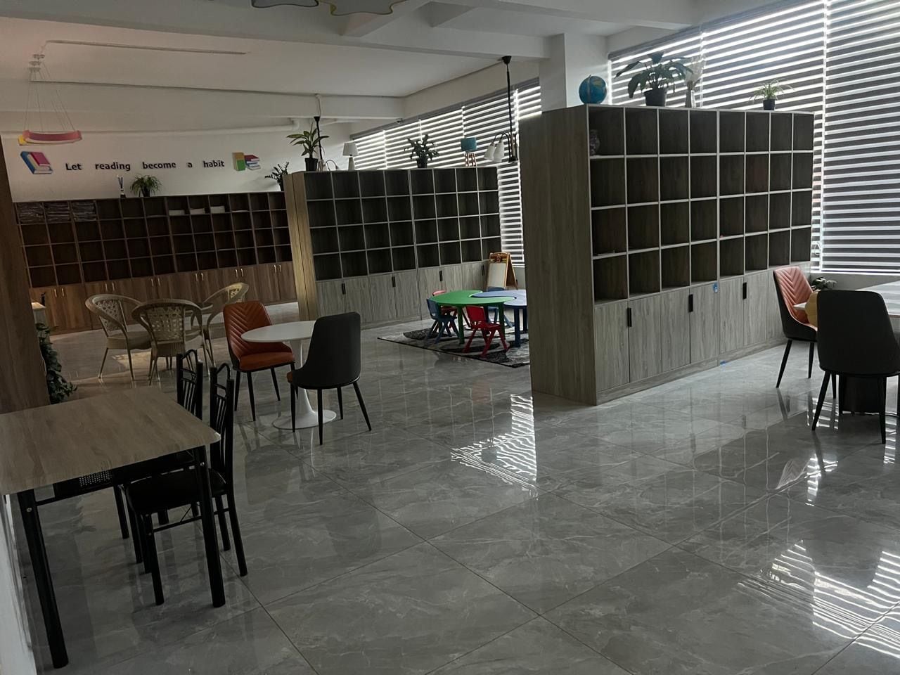 Una biblioteca moderna con una distribución limpia, que presenta una larga mesa con sillas, una estantería y un área de lectura con una mesa pequeña y sillas.