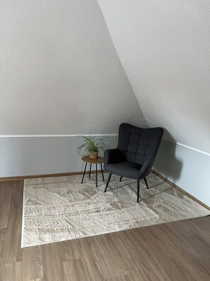 Ein Eckzimmer mit grauem Polsterstuhl, einem kleinen runden Tisch mit Topfpflanze und einem gemusterten Teppich auf Holzdielen.