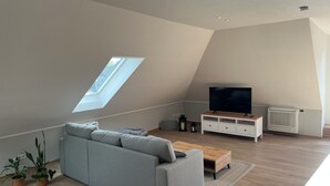 Quarto moderno estilo loft com teto inclinado, claraboia, sofá cinza, mesa de centro de madeira, TV em suporte e radiador.