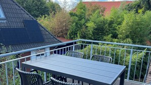 Um deck de madeira com mesa e cadeiras, rodeado por um guarda-corpo de metal, com vista para um jardim e um edifício com telhado vermelho.