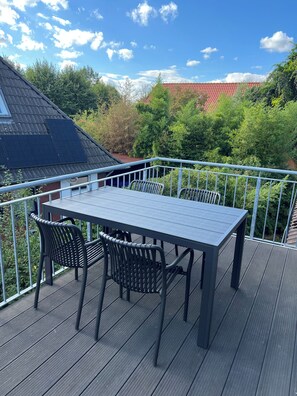 Een houten terras met een tafel en stoelen, omgeven door een metalen hek, met uitzicht op een tuin en een gebouw met een rood dak.