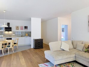 Eine moderne Küche mit weißen Schränken, ein Essbereich mit Holztisch und -stühlen sowie ein Wohnzimmer mit beigefarbenem Sofa und farbenfrohem Teppich.