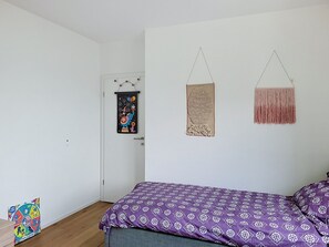 Quarto com colcha roxa estampada, porta com arte colorida e dois objetos decorativos pendurados na parede.