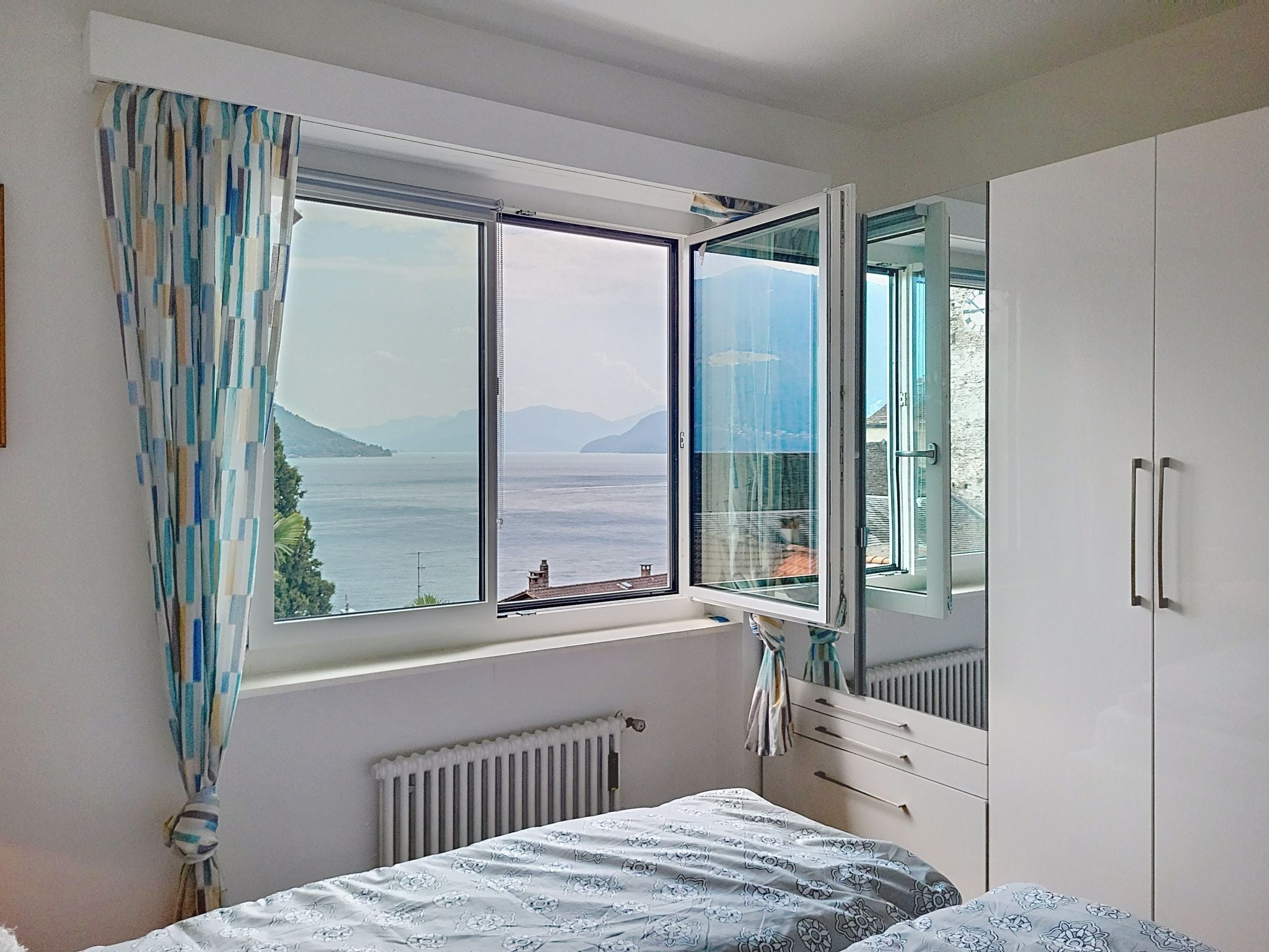 A bedroom with a large window, a bed with patterned bedding, a radiator, and a wardrobe.