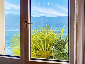 A window with a view of a mountain range and a body of water, with a palm plant in the foreground. - Casa Matani (Seezugang) by Interhome (Ranzo)