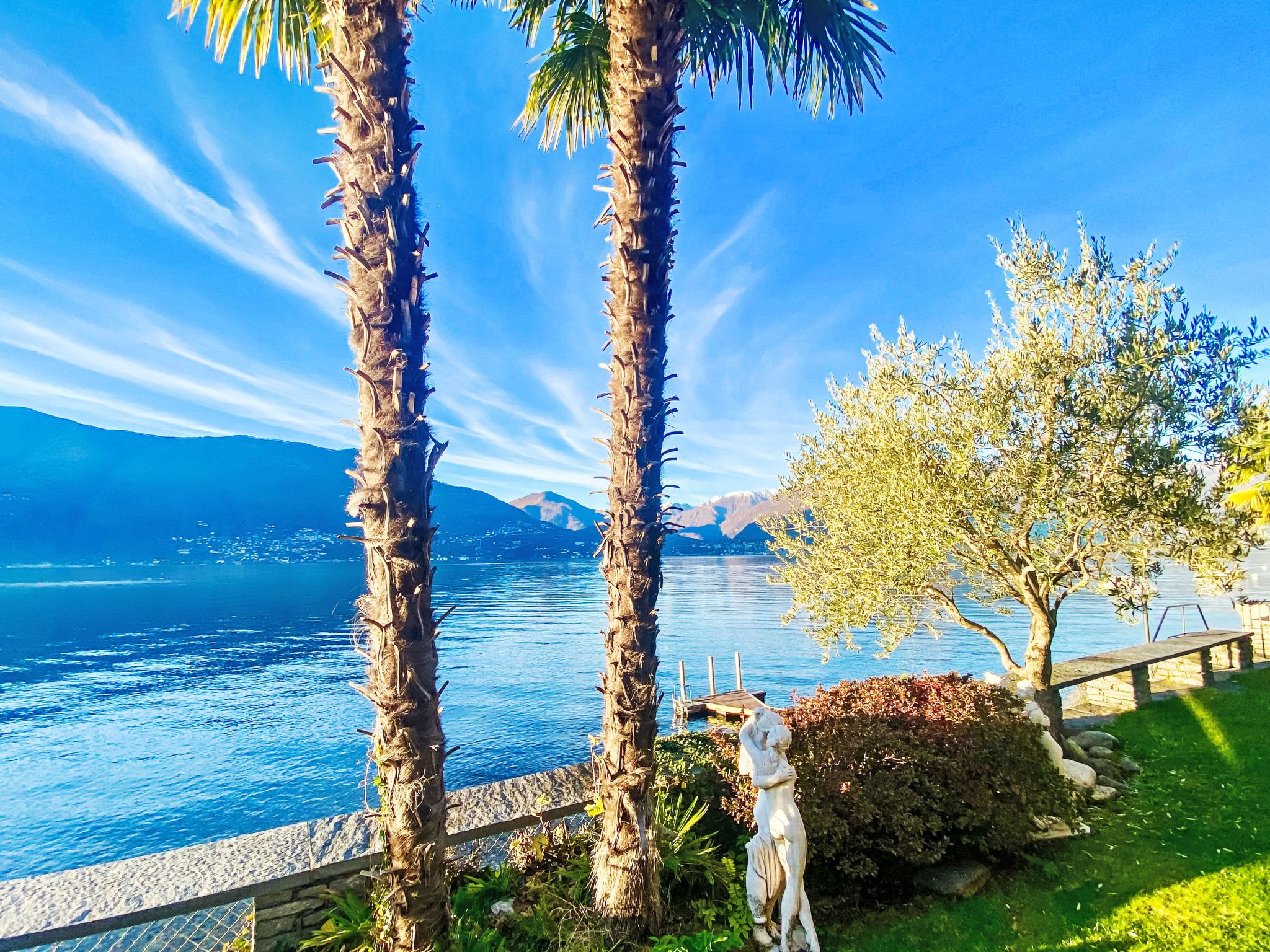 A coastal landscape with palm trees, a statue, and a view of the sea and mountains.