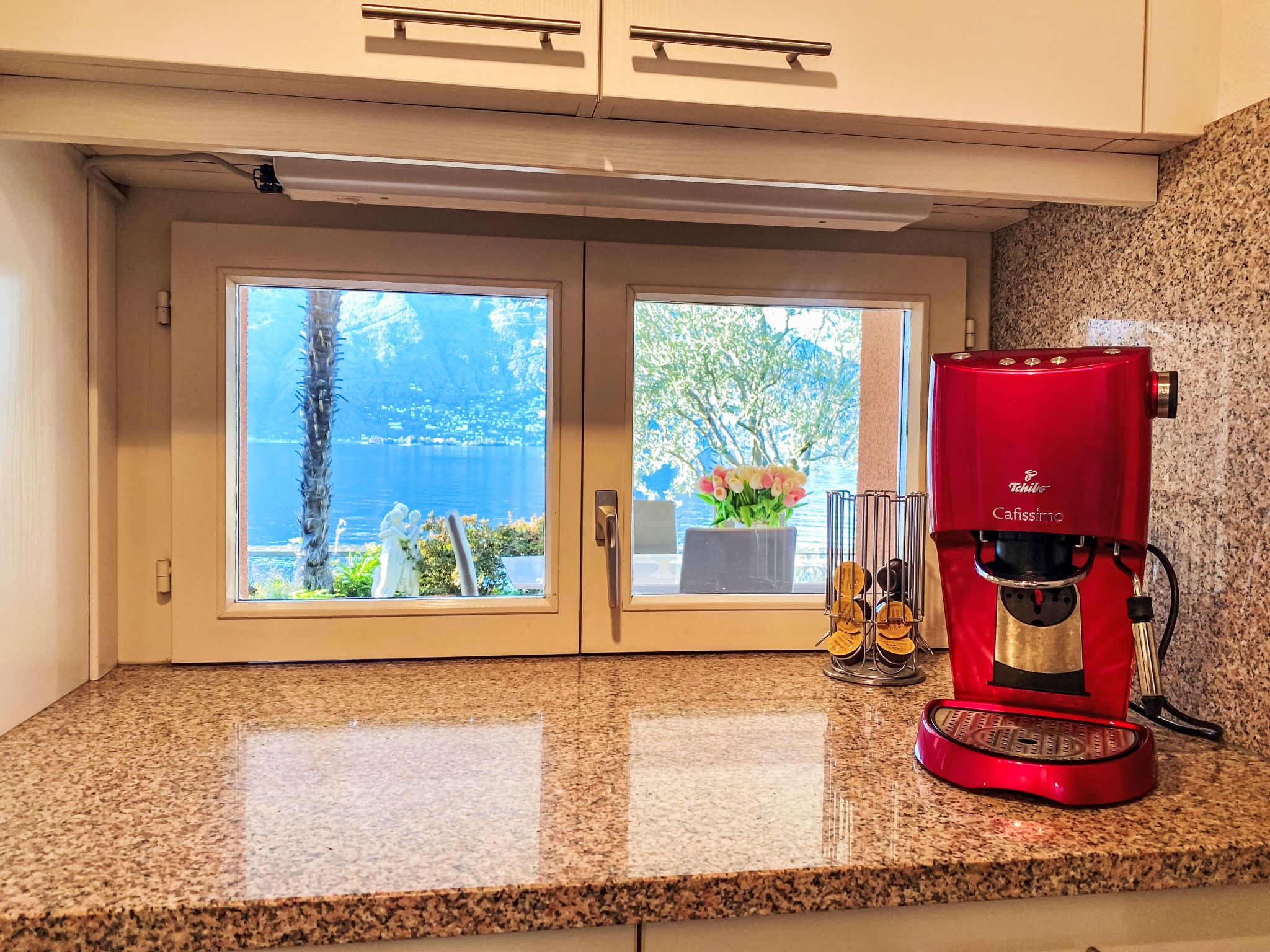 A kitchen with a red coffee machine, a window with a view of the sea, and a granite countertop.