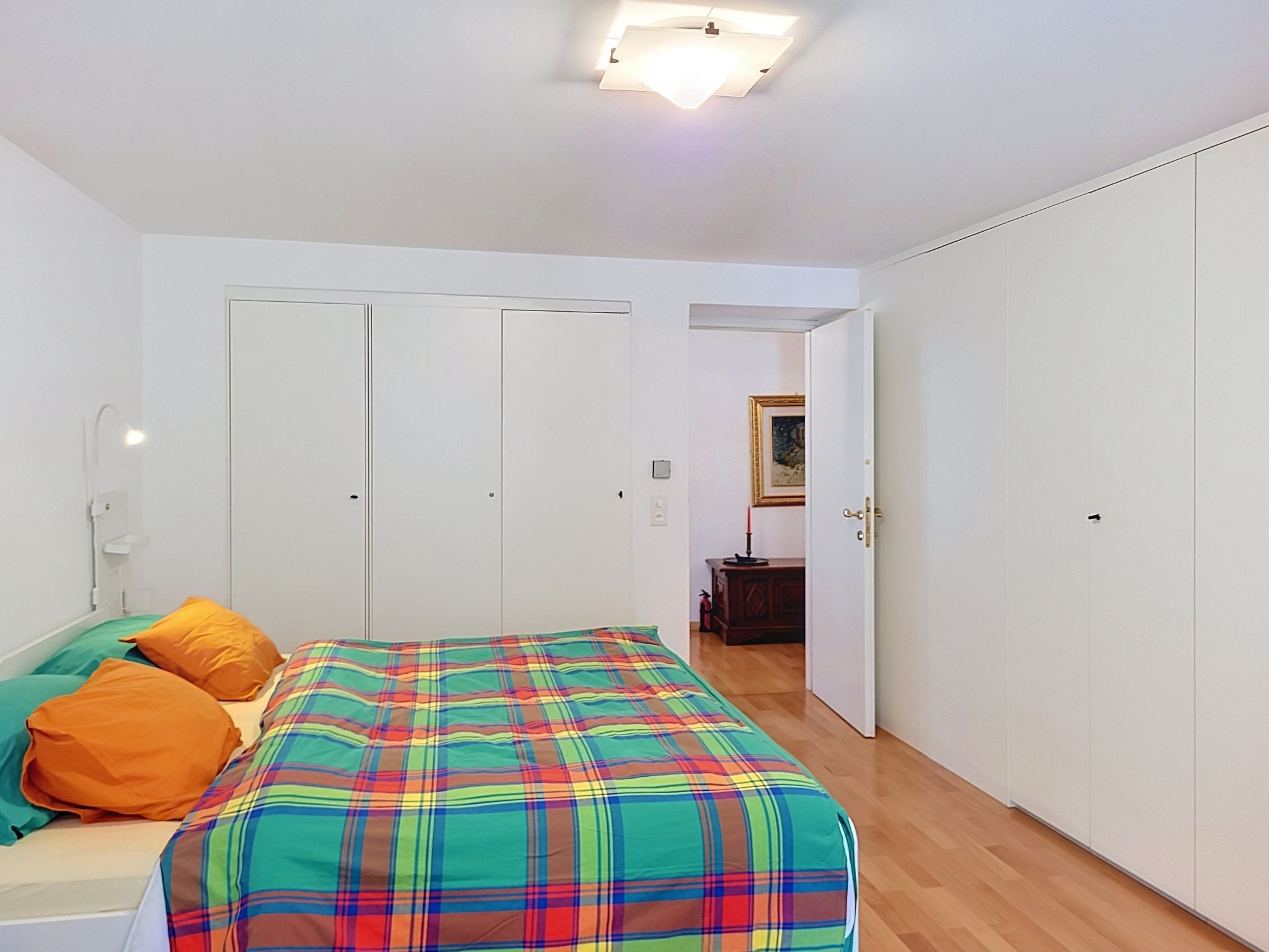 Ein Schlafzimmer mit einem Bett, zwei Kissen, einem Nachttisch, einer Stehlampe, einem Kleiderschrank und einer Tür zu einem weiteren Raum.