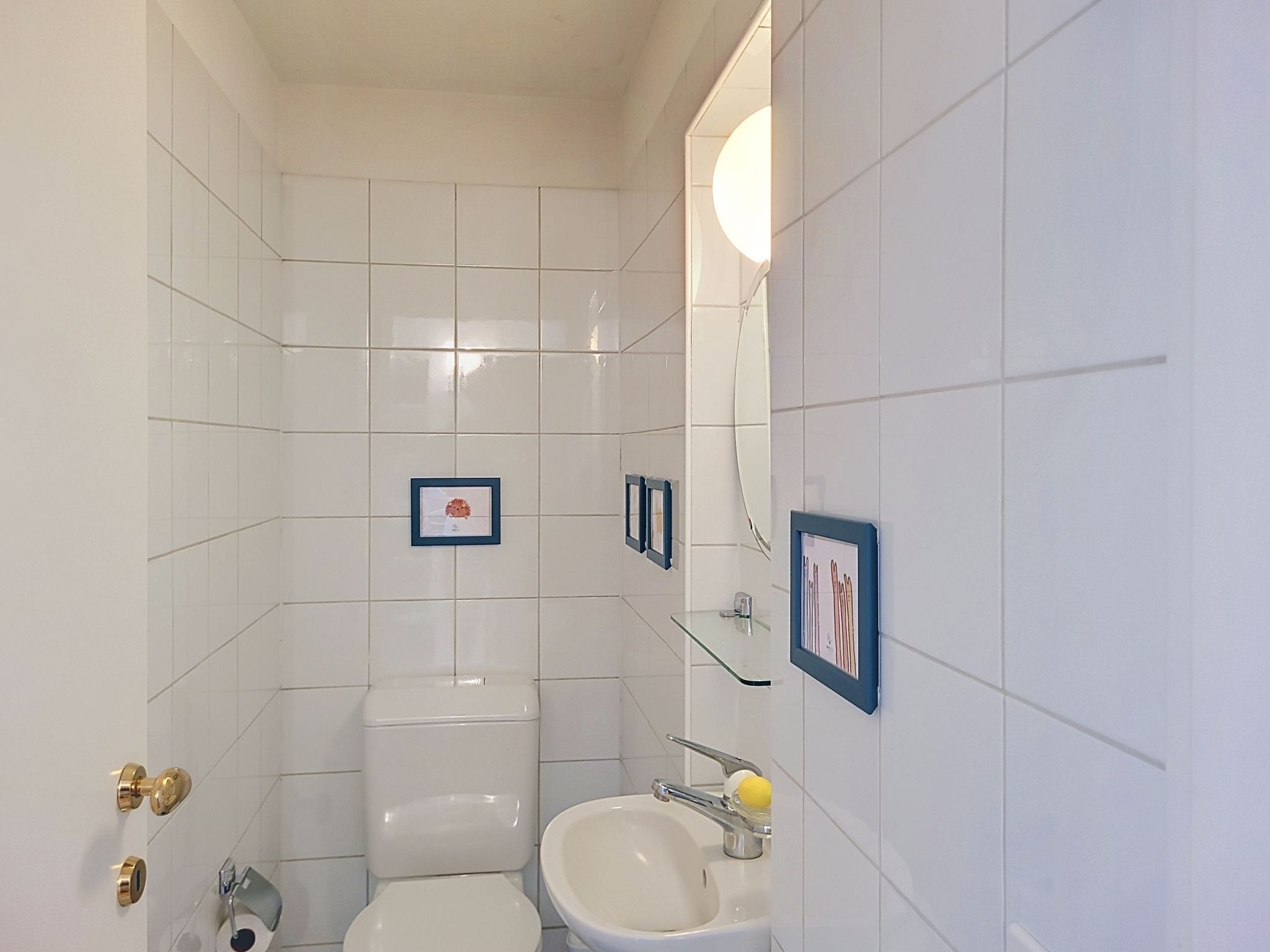 Une salle de bain avec des murs carrelés de blanc, des toilettes, un lavabo avec un miroir et des tableaux encadrés accrochés au mur.
