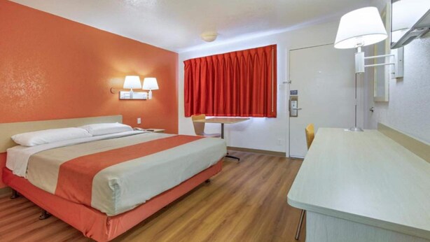Quarto de hotel com cama, escrivaninha, cadeira, janela com cortinas vermelhas e luminária na parede.